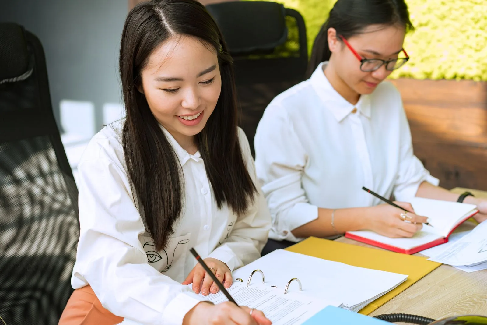 Two Asian payroll staff writing notes from a binder of salary records to work out Personal Income Tax withholding